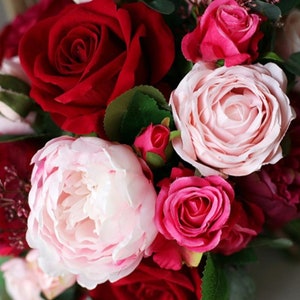 May include: A bouquet of artificial flowers in shades of pink and red. The bouquet includes roses, peonies, and smaller pink flowers.