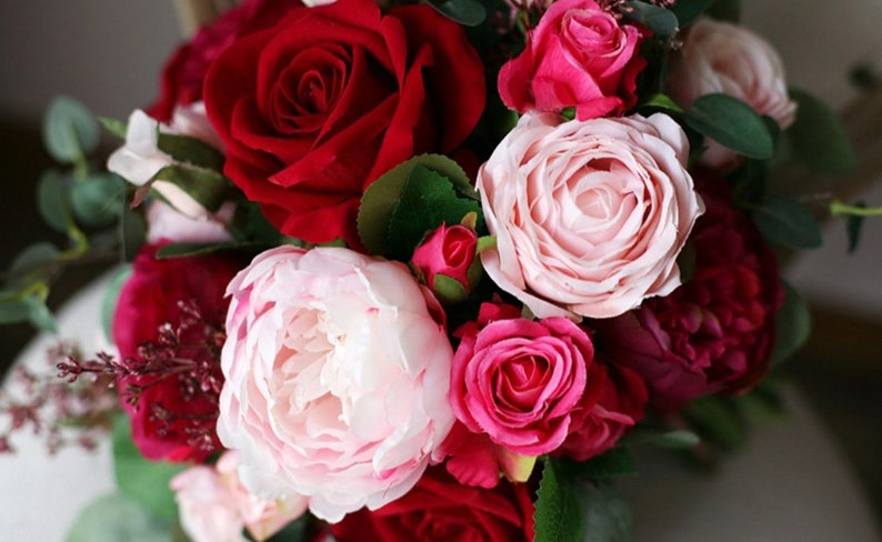 May include: A bouquet of artificial flowers in shades of pink and red. The bouquet includes roses, peonies, and smaller pink flowers.