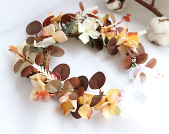 May include: A floral crown with brown and yellow flowers and green leaves. The crown is tied with a white ribbon.