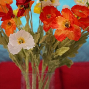 May include: A bouquet of artificial poppies in a glass vase. The flowers are red, orange, white, and yellow. The vase is clear glass and has a textured surface.