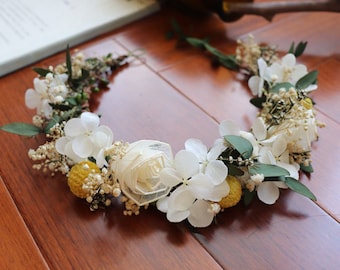May include: A floral crown featuring white hydrangea, small yellow accents, and greenery. The crown is arranged in a circular shape, perfect for weddings or special occasions. The flowers are set against a wooden surface.
