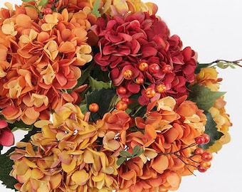Könnte beinhalten: Ein Blumenarrangement mit künstlichen Hortensienblüten in den Farben gebranntes Orange, Goldgelb und tiefem Burgunderrot. Der Strauß ist mit kleinen, runden orangefarbenen und roten Beeren sowie grünen Blättern an dünnen Stielen verziert.