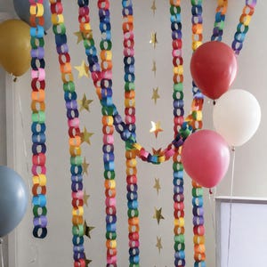 Puede incluir: Una escena festiva con guirnaldas de cadenas de papel de colores del arcoíris, decoraciones de estrellas doradas y globos rojos, rosas, dorados y azules. Las decoraciones están dispuestas sobre un fondo blanco.