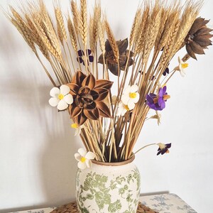 Könnte beinhalten: Eine weiße Keramikvase mit grünem Blumenmuster, gefüllt mit getrockneten Weizenhalmen, braunen Sternanis-Kapseln und weißen und violetten Blumen.