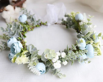 May include: A light blue floral crown with white flowers and greenery. The crown is made of artificial flowers and is perfect for a wedding or other special occasion.