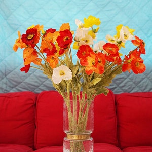 May include: A glass vase with a gold band around the middle, filled with a bouquet of orange, yellow, and white poppy flowers. The vase is sitting on a table with a patterned tablecloth.