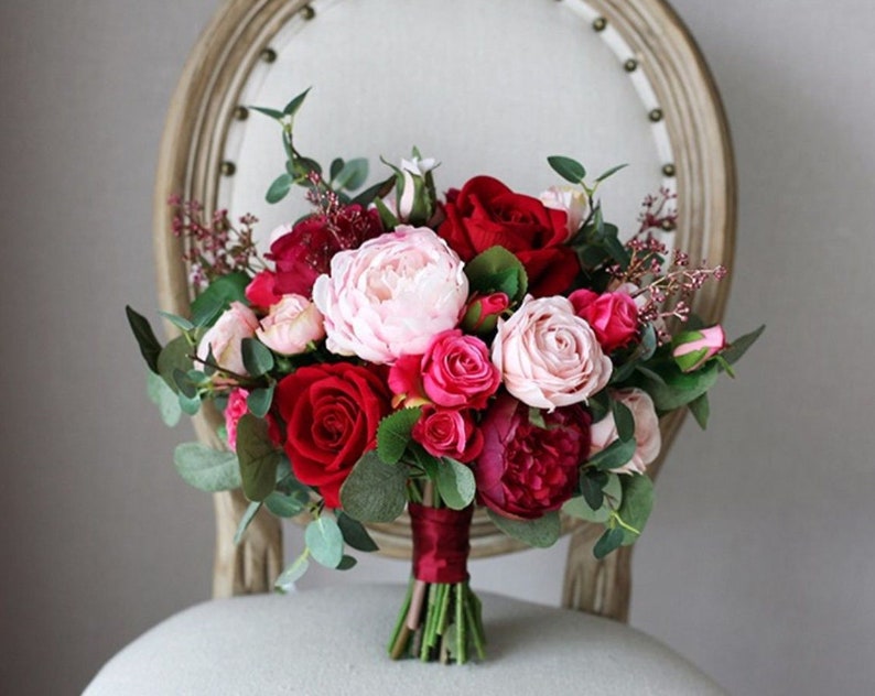 May include: A bridal bouquet with red, pink, and white roses, peonies, and greenery. The bouquet is tied with a burgundy ribbon.