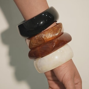May include: A stack of four wide bangle bracelets worn on the wrist. The set includes one solid glossy black bangle, two marbled brown bangles with varying amber and mahogany tones, and one smooth off-white cream bangle with a subtle marbled texture.