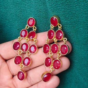 May include: A pair of gold-toned chandelier earrings with oval-shaped, deep red gemstones. The earrings feature multiple tiers of gemstones, creating a cascading effect. The gemstones are set in gold bezels.