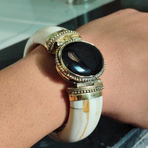 May include: A hinged bangle bracelet featuring two curved off-white segments with subtle tan striations. The centre is adorned with a large, oval, polished black stone set in a gold-toned metal frame with a beaded border and a hinged clasp mechanism.