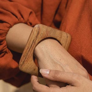以下が含まれることがあります： 丸みを帯びたエッジを持つ正方形の木製ブレスレット。ブレスレットは明るい茶色の木材で作られており、手首に着用されています。その人は錆色の長袖シャツを着ています。