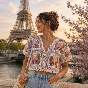 May include: A lilac, pink, and yellow crochet butterfly cardigan with denim shorts. The short-sleeved top has a button-up front and a scalloped edge. A straw tote bag hangs from the shoulder. The Eiffel Tower is in the background.