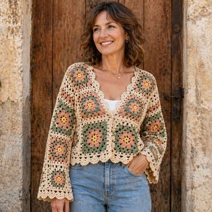 Può includere: Un cardigan all'uncinetto beige, verde e rosa con un motivo floreale. Il cardigan a maniche lunghe ha uno scollo e bordi smerlati. La donna indossa un top bianco e jeans blu.