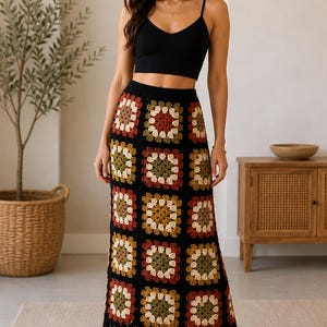 Op de afbeelding: Zwarte gehaakte rok met een kleurrijk bloemenpatroon, gecombineerd met een zwarte crop top. De rok heeft een herhalend vierkant ontwerp in rood, geel, groen en crème. De outfit wordt afgemaakt met zwarte sandalen.