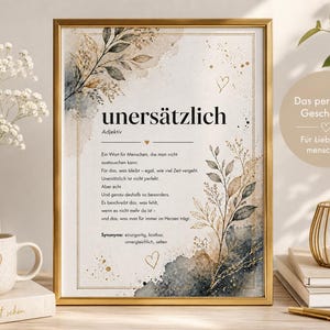 Puede incluir: Una impresión enmarcada con la palabra alemana "unersätzlich" en una fuente decorativa, con acentos de estilo acuarela e ilustraciones botánicas. La impresión está en un marco dorado, con una taza de café y un jarrón en primer plano.