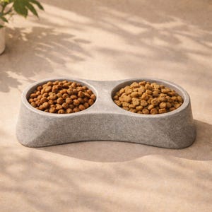 May include: A gray, double pet bowl filled with dry pet food. The bowl has a curved design with two circular compartments. The food is a light brown color, and the bowl is made of a stone-like material.