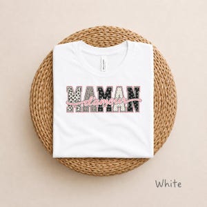 Peut inclure: Un t-shirt blanc à manches courtes avec le texte MAMAN d'amour en lettres capitales ornées de motifs noirs et blancs variés comme des léopards, des nœuds et des étoiles, souligné par un contour rose et une écriture cursive rose.