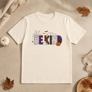 May include: A cream-colored short-sleeve t-shirt featuring the text BE KIND in a Halloween-themed font. The letters are decorated with spiderwebs, orange and black plaid, stars, and ghosts. A small orange pumpkin is positioned next to the letter D.