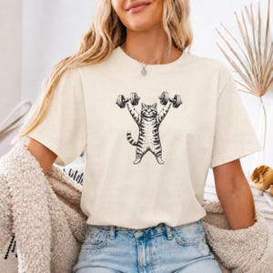 Könnte beinhalten: Ein cremefarbenes Kurzarm-T-Shirt mit einer schwarzen Grafik im Tintenstil, die eine getigerte Katze zeigt, welche zwei Hanteln über ihren Kopf hebt. Die Katze ist stehend mit geringeltem Schwanz dargestellt und weist ein detailliertes Design auf.