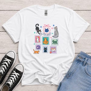 Può includere: Una t-shirt bianca a maniche corte con una stampa grafica colorata di vari gatti in cornici a forma di francobollo, tra cui una testa di gatto blu in una cornice con fiocco rosa con il testo CAT LOVER, circondata da stelle e gatti.