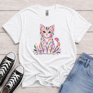 May include: A white short-sleeved t-shirt featuring a central graphic of a sitting cat rendered in colorful, painterly brushstrokes of pink, blue, yellow, and green. The cat is surrounded by stylized leaf shapes at its base, creating a vibrant, artistic design on the front of the garment.