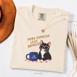 May include: An ivory t-shirt featuring a graphic of a black and white cat wearing pink heart-shaped sunglasses next to a blue coffee mug. The text reads MORE ESPRESSO LESS DEPRESSO and I LOVE COFFEE on the mug. The shirt has a Comfort Colors label.