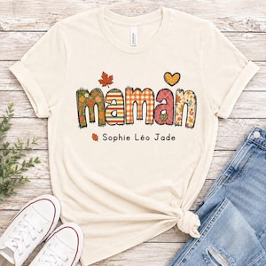 Może przedstawiać: Kremowy t-shirt z krótkim rękawem z napisem "maman" wykonanym czcionką w stylu patchwork z jesiennymi wzorami, w tym kratką, paskami i liśćmi. Pod główną grafiką znajduje się małe serce oraz tekst "Sophie Léo Jade".