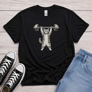 Può includere: Una t-shirt nera a maniche corte con una stampa grafica di un gatto tigrato che solleva un bilanciere sopra la testa. Il gatto è raffigurato in uno stile illustrativo chiaro e dettagliato, centrato sul petto della maglietta.