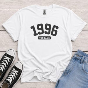 Peut inclure: Un t-shirt blanc à manches courtes arborant le texte noir 1996 dans une police universitaire grasse, avec le mot VINTAGE imprimé dans un petit rectangle noir juste en dessous des chiffres, centré sur la poitrine du vêtement.