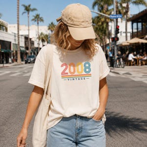 May include: A beige baseball cap and a cream-colored t-shirt featuring the text 2008 VINTAGE in a multi-colored, striped, retro-style font. The shirt is paired with light blue denim jeans and a matching cream-colored canvas tote bag.