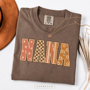 Puede incluir: Una camiseta marrón de manga corta con cuello redondo y etiqueta Comfort Colors. En la parte delantera aparece la palabra NANA en letras grandes de bloque, cada una con un patrón diferente: estrellas, cuadros, rayas y flores.