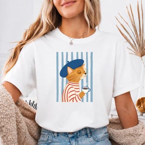 Può includere: Una t-shirt bianca con una stampa grafica di un gatto soriano arancione che indossa un basco blu, occhiali rotondi e una maglia a righe rosse e bianche. Il gatto tiene in mano una piccola tazza da caffè bianca su uno sfondo a righe blu.