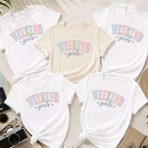 Peut inclure: Quatre t-shirts à manches courtes, trois blancs et un beige, arborant chacun le texte WEEKEND dans une police à carreaux multicolores et le mot girls en écriture cursive bleue, agrémenté de deux petits cœurs bleus de chaque côté.