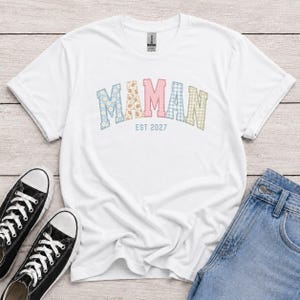 Può includere: Una t-shirt bianca a maniche corte con la parola MAMAN in un carattere a blocchi multicolore con motivi floreali e a quadretti vichy. Sotto la parola, il testo EST 2027 è stampato in un semplice carattere blu. Ha un girocollo.