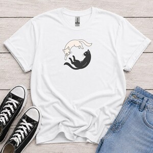 Könnte beinhalten: Ein weißes Kurzarm-T-Shirt mit einer zentralen Grafik, die eine cremefarbene Katze und eine schwarze Katze in einer kreisförmigen Yin-Yang-Anordnung zeigt. Die Katzen sind mit einfachen Umrissen und minimalistischen Merkmalen dargestellt.