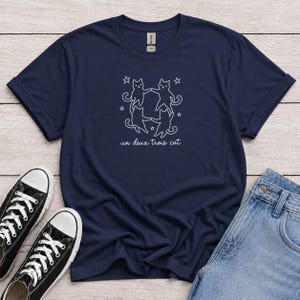 Peut inclure: Un t-shirt bleu marine à manches courtes avec un motif linéaire blanc représentant quatre chats dansant en cercle entourés de petites étoiles. Sous les chats, le texte un deux trois cat est écrit en police cursive blanche. Col rond.