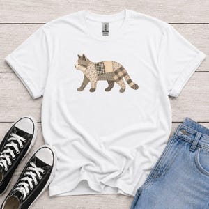 Puede incluir: Una camiseta blanca de manga corta con un gráfico de un gato compuesto por varios patrones de patchwork en tonos marrón y beige, incluyendo texturas de cuadros, flores y colores sólidos. El gato se muestra de perfil en posición de caminar.