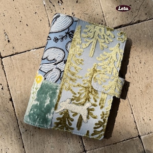 Może przedstawiać: Jasnoniebieskie i złote haftowane etui na telefon z motywem lasu i królika. Etui posiada zapięcie na klapę i jest wykonane z miękkiej, teksturowanej tkaniny. Etui jest ozdobione wzorem kwiatowym i liściastym.