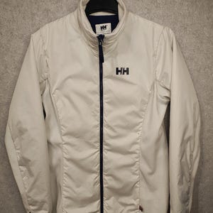May include: A white zip-up jacket featuring a high collar and a dark blue front zipper. The jacket displays the HH logo in dark blue on the left chest and on the inner neck label. It has long sleeves and a smooth, solid-colored outer shell fabric.