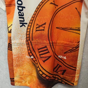 Puede incluir: Maillot de ciclismo naranja y crema con un dise&ntilde;o de reloj y n&uacute;meros romanos. El maillot tiene tres bolsillos traseros y el texto "rabobank" y "agu".