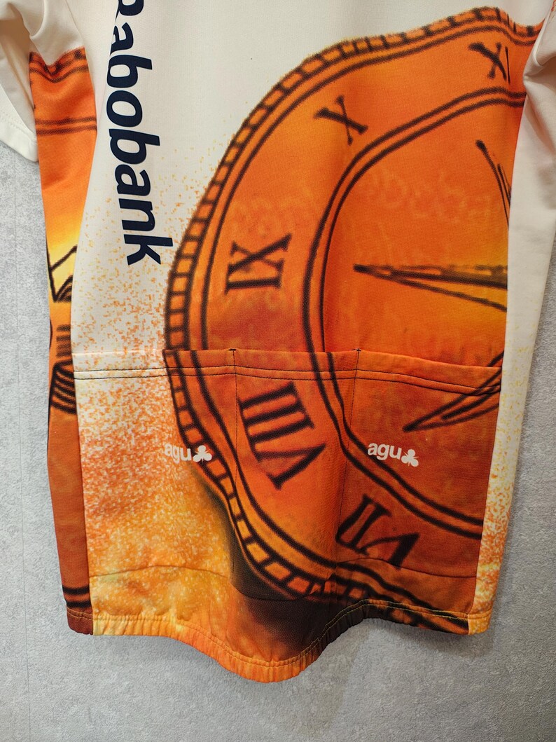 Puede incluir: Maillot de ciclismo naranja y crema con un dise&ntilde;o de reloj y n&uacute;meros romanos. El maillot tiene tres bolsillos traseros y el texto "rabobank" y "agu".