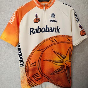 Puede incluir: Maillot de ciclismo con cuerpo blanco y mangas naranjas. El maillot presenta el logo "Rabobank" en azul marino, un dise&ntilde;o de reloj y el logo "agu". El maillot tiene una cremallera corta y est&aacute; colgado de una percha negra.