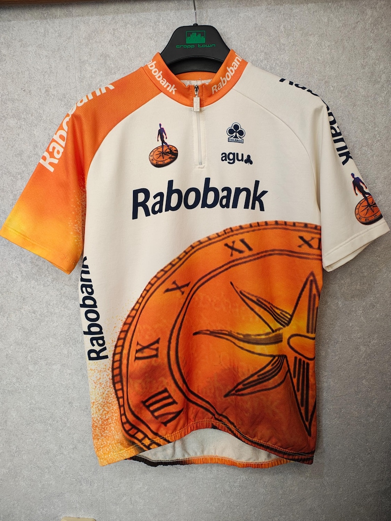 Puede incluir: Maillot de ciclismo con cuerpo blanco y mangas naranjas. El maillot presenta el logo "Rabobank" en azul marino, un dise&ntilde;o de reloj y el logo "agu". El maillot tiene una cremallera corta y est&aacute; colgado de una percha negra.