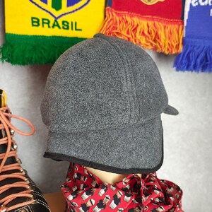 May include: A gray fleece cap with a small brim and black trim. The cap is displayed on a mannequin head, with a red scarf featuring a repeating pattern of figures. In the background are colorful soccer scarves.