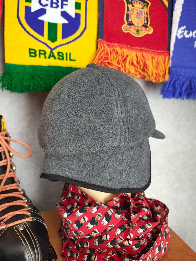 May include: A gray fleece cap with a small brim and black trim. The cap is displayed on a mannequin head, with a red scarf featuring a repeating pattern of figures. In the background are colorful soccer scarves.