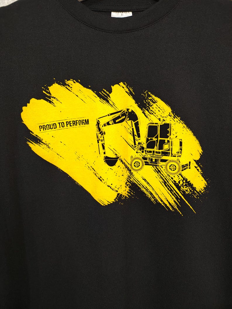 K&ouml;nnte beinhalten: Schwarzes Sweatshirt mit gelber Bagger-Grafik und dem Text "PROUD TO PERFORM" auf einem gelben Pinselstrich. Der Bagger ist eine detaillierte Silhouette. Das Design ist auff&auml;llig und ansprechend.