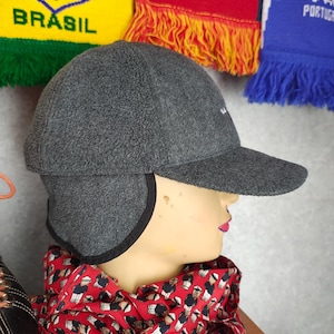 May include: A gray fleece cap with ear flaps and a curved brim. The cap has a small white logo on the front. A red scarf with a repeating pattern of figures is wrapped around the neck of a mannequin. Soccer scarves are in the background.