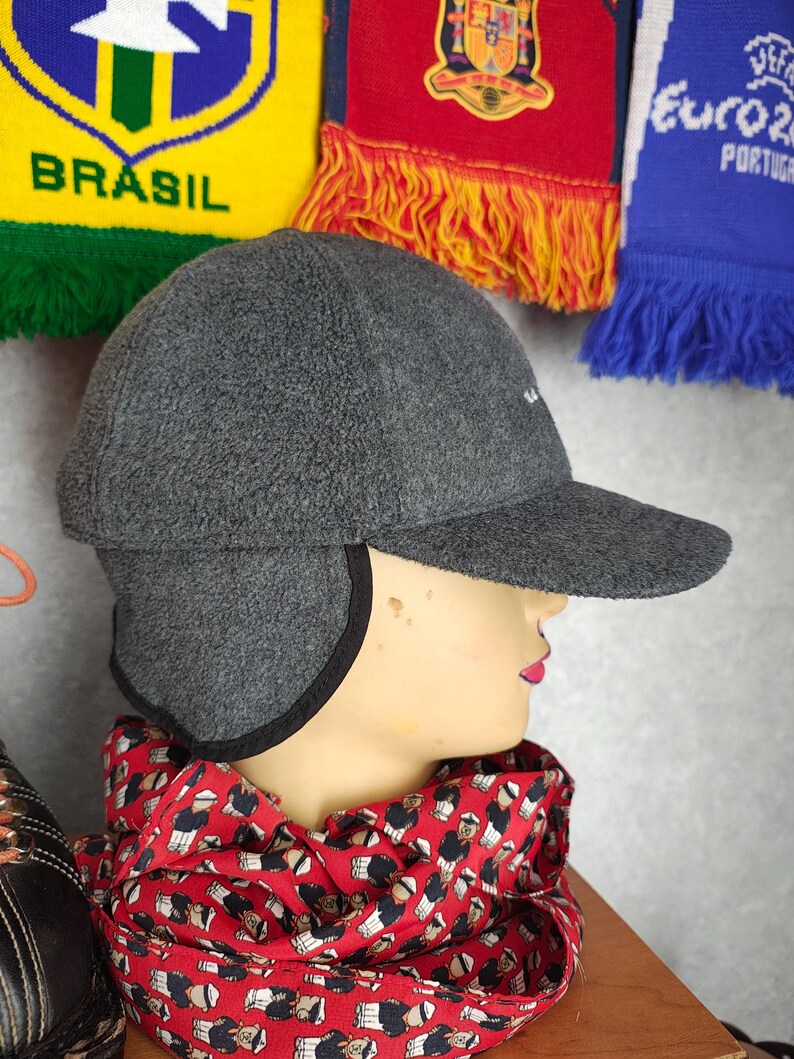 May include: A gray fleece cap with ear flaps and a curved brim. The cap has a small white logo on the front. A red scarf with a repeating pattern of figures is wrapped around the neck of a mannequin. Soccer scarves are in the background.