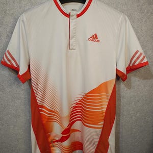 Może przedstawiać: Biała koszulka sportowa z pomarańczowymi i czerwonymi akcentami. Posiada czerwone logo Adidas na piersi. Koszulka ma kołnierzyk mandaryński i krótkie rękawy z czerwonymi paskami. Projekt zawiera pomarańczowe i czerwone zakrzywione linie.