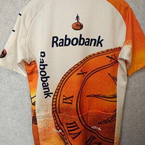 Puede incluir: Maillot de ciclismo color crema con detalles en naranja y el logo "Rabobank". El maillot presenta un gran dise&ntilde;o de reloj y el texto "de lage landen". La espalda tiene tres bolsillos.